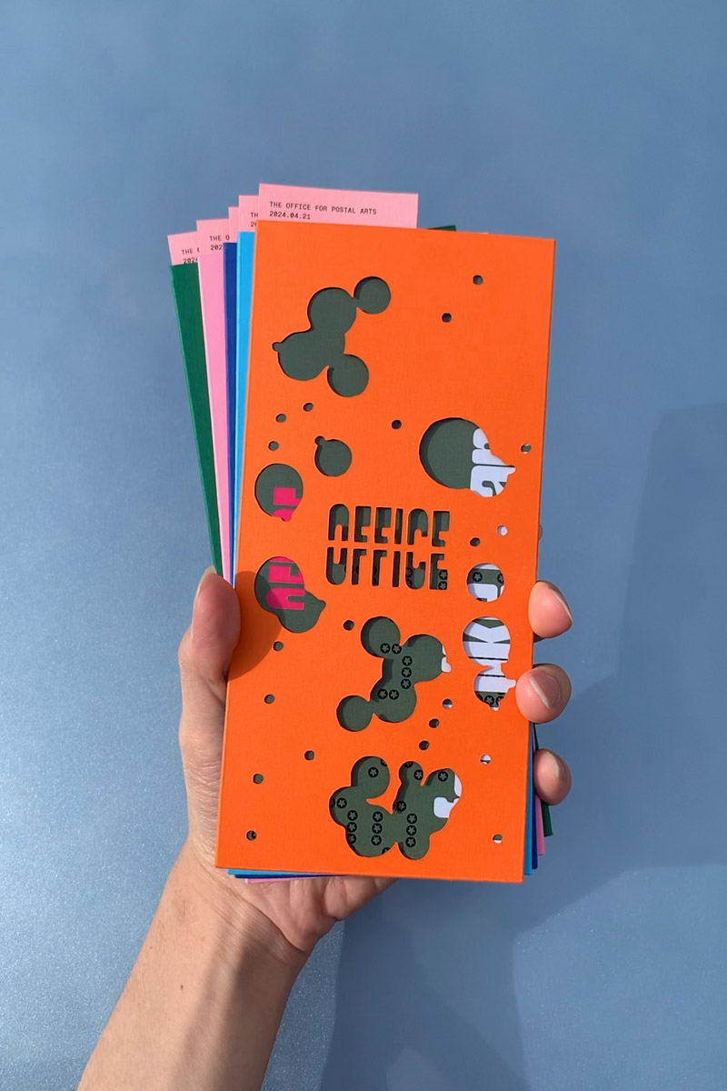 Photo of several multi-coloured copies of the Office for Postal Arts publication fanned out, held by a hand.
